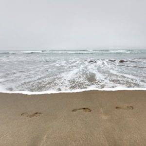 Three footprints are clearly imprinted on a beach. White foam almost reaches them, and a crest is visible in the distance under a gray sky.