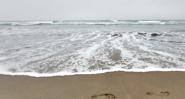 Three footprints are clearly imprinted on a beach. White foam almost reaches them, and a crest is visible in the distance under a gray sky.