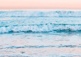 A line of pastel blue waves rolls towards the camera against a pink dawn sky