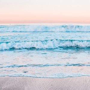 A line of pastel blue waves rolls towards the camera against a pink dawn sky