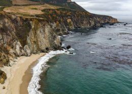 Dark patches of kelp stand out against the teal of the Big Sur coast. Highway 101 is visible on the clifftops