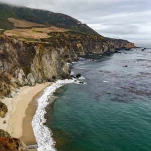 Dark patches of kelp stand out against the teal of the Big Sur coast. Highway 101 is visible on the clifftops