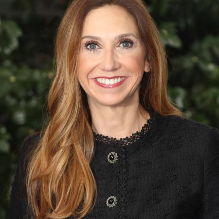 Photograph of a white woman with auburn hair smiling at the camera. She wears a black blouse with ornate buttons.