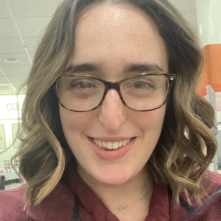 A young white woman with glasses and a nostril piercing smiles at the camera.