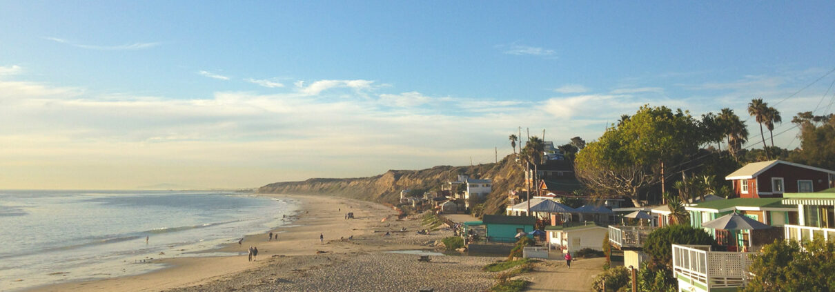 A wide beach during the golden hour just before sunset, with houses in the foreground and cliffs stretching away to the north