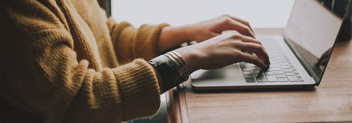 A seated person wearing a yellow sweater and bracelets typing on a laptop