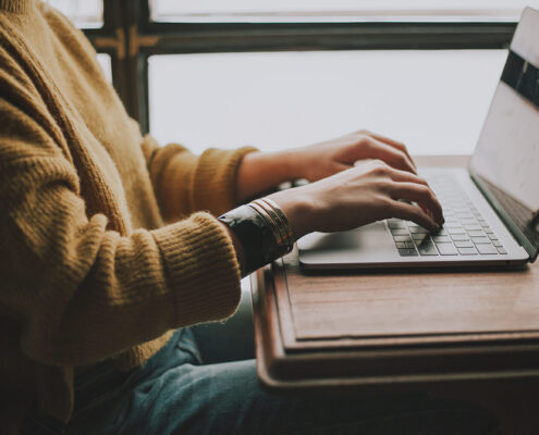 A seated person wearing a yellow sweater and bracelets typing on a laptop