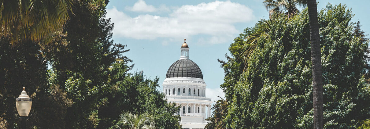 View of the Sacramento Capitol Building down a street lined with trees, with a blue sky and light clouds behind the dome