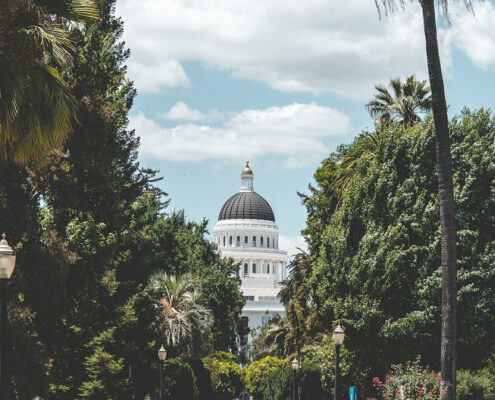 View of the Sacramento Capitol Building down a street lined with trees, with a blue sky and light clouds behind the dome