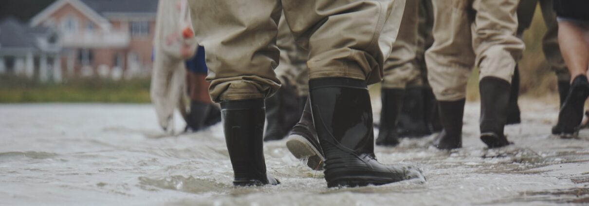 Several pairs of black rubber boots and brown waterproof pants face away from the viewer, standing ankle-deep in floodwaters