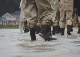 Several pairs of black rubber boots and brown waterproof pants face away from the viewer, standing ankle-deep in floodwaters