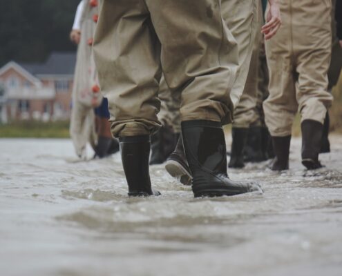Several pairs of black rubber boots and brown waterproof pants face away from the viewer, standing ankle-deep in floodwaters