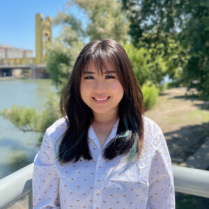 A young Asian woman with dark hair smiles at the camera. The background is a sunny view of the Sacramento River, with the yellow Tower Bridge visible among the foliage of the background.