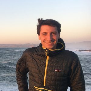 Photo of a young man with short hair and a dark jacket standing in front of the ocean at sunset