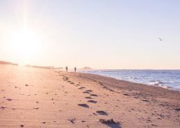 Photo of two silhouetted people walking along a beach in the morning, with the sun shining brightly into the camera. Their footprints are clearly visible on the sand of the beach.