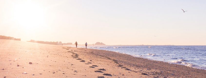 Photo of two silhouetted people walking along a beach in the morning, with the sun shining brightly into the camera. Their footprints are clearly visible on the sand of the beach.