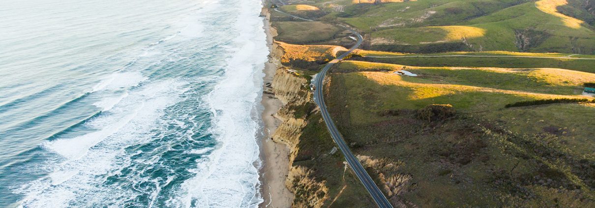 Aerial photograph of CA Highway 101 and the coastline. Waves are visible in the water and green hills roll away to the east.