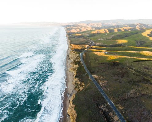 Aerial photograph of CA Highway 101 and the coastline. Waves are visible in the water and green hills roll away to the east.