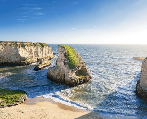 Cliffs and a large rock rise out of the shallow water of a beach at Shark Fin Cove. Late afternoon sunlight casts long shadows.