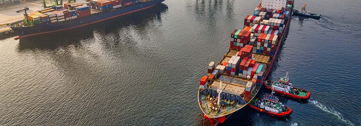 Three tugboats push a container ship in towards cranes in a port.