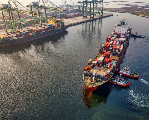 Three tugboats push a container ship in towards cranes in a port.