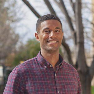 Photograph of a young Black man wearing a plaid shirt smiling at the camera