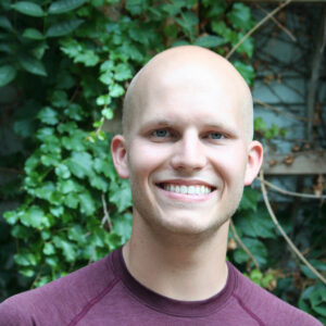 A young bald white man smiles at the camera. He is wearing a maroon shirt and stands against a leafy green background.