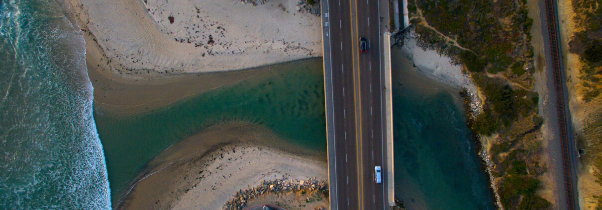An aerial photograph of Cardiff Beach in Encinitas. A river flows out under a bridge, while a wide sandy beach curls under it and waves break beyond.