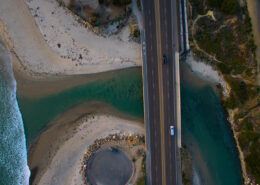 An aerial photograph of Cardiff Beach in Encinitas. A river flows out under a bridge, while a wide sandy beach curls under it and waves break beyond.