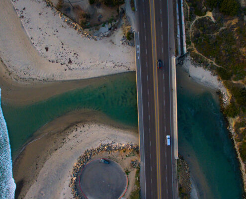 An aerial photograph of Cardiff Beach in Encinitas. A river flows out under a bridge, while a wide sandy beach curls under it and waves break beyond.