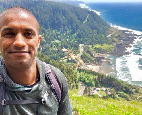 Photograph of a young Black man smiling at the camera. He stands on a hillside overlooking a beach and further hills.