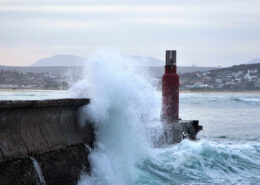 Waves crash dramatically against a breakwater. A town is visible in the background, nestled into the low hills by the beach.