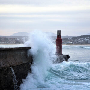 Waves crash dramatically against a breakwater. A town is visible in the background, nestled into the low hills by the beach.