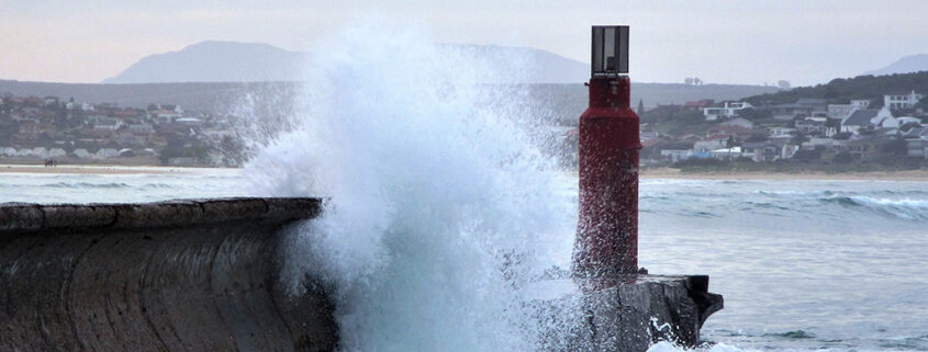 Waves crash dramatically against a breakwater. A town is visible in the background, nestled into the low hills by the beach.