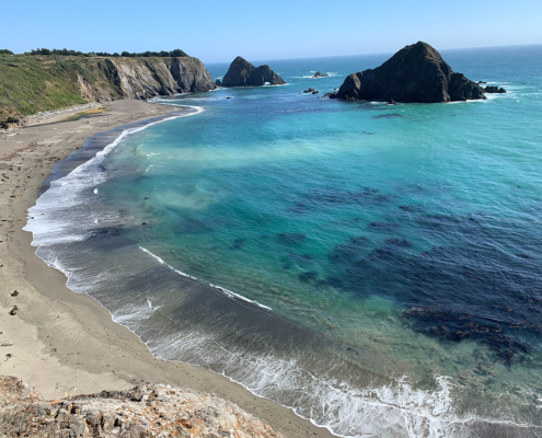 Cliffs and a beach on a clear day along the Mendocino coast. Dark patches of kelp are visible in the turquoise water.