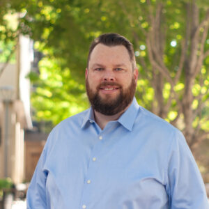 A dark-haired white man in a blue shirt smiles at the camera.