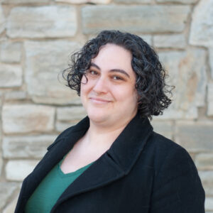 A person with dark curly hair and a dark coat smiles at the camera.