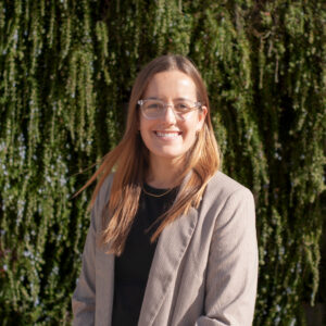 A young white woman with dark blonde hair smiles at the camera. She wears a brown blazer and glasses.