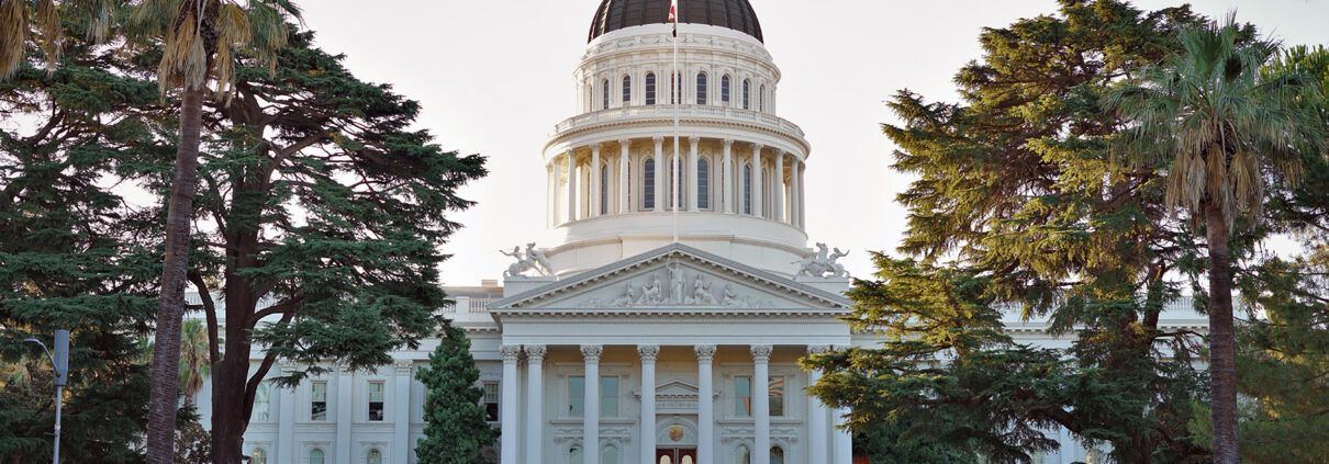 Photograph of the California Capitol Building from in front. The sky in the background is pale in predawn and there are trees to either side of the cupola.