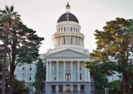 Photograph of the California Capitol Building from in front. The sky in the background is pale in predawn and there are trees to either side of the cupola.
