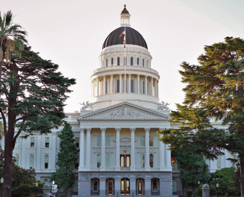 Photograph of the California Capitol Building from in front. The sky in the background is pale in predawn and there are trees to either side of the cupola.