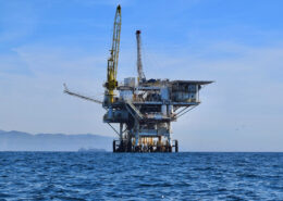 Photograph of a large oil and gas rig off the coast of Santa Barbara. The Coastal Range is visible in the background above the waves.