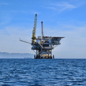 Photograph of a large oil and gas rig off the coast of Santa Barbara. The Coastal Range is visible in the background above the waves.