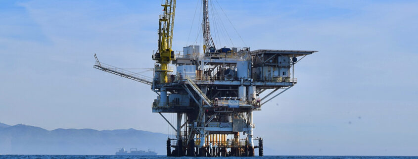 Photograph of a large oil and gas rig off the coast of Santa Barbara. The Coastal Range is visible in the background above the waves.