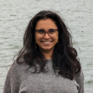 Headshot of a young Desi woman smiling at the camera in front of gray waves. She wears a gray sweater and glasses, and has dark curly hair.