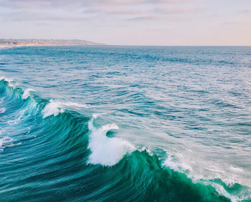 A low wave cresting towards a distant shore. A long line of whitecaps is visible, as are dunes along the horizon.