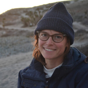 A young white woman wearing glasses and a navy blue beanie smiles a the camera