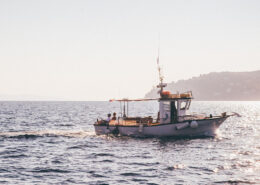 Photograph of a small fishing boat on calm waters. A low headland is visible in the distance, and two small figures sit on the back of the boat.