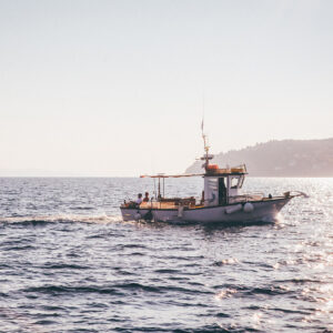 Photograph of a small fishing boat on calm waters. A low headland is visible in the distance, and two small figures sit on the back of the boat.