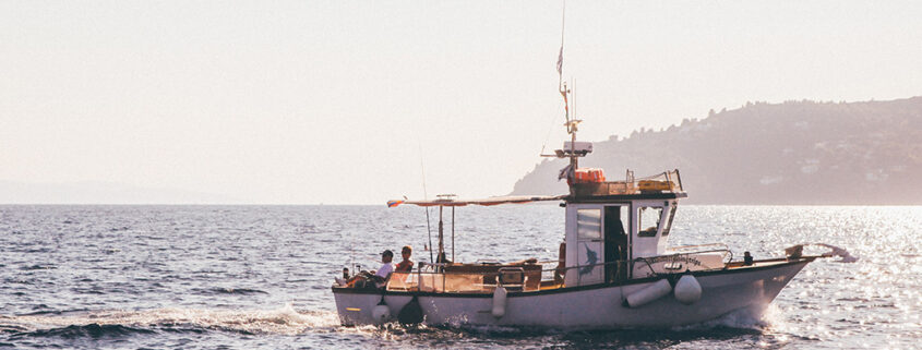 Photograph of a small fishing boat on calm waters. A low headland is visible in the distance, and two small figures sit on the back of the boat.